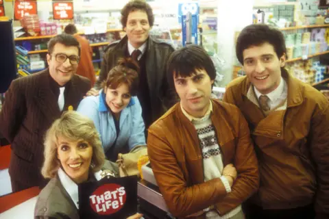 The cast of That's Life gathered around a supermarket checkout in 1983. The cast consists of four men and two women, including Bill Buckley who has black short hair and a brown zip-up jacket. Also in the photo is lead presenter Esther Ranzen, who has blonde hair and is holding up a clip board.