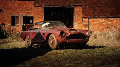 Crossley Motorsport A dilapidated red Aston Martin from 1960 parked on some grass outside a red-brick garage.