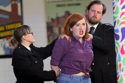 Sartorial Pictures A scene from Carmen. Two security people - one man and one woman are holding a woman, who has long red hair and is wearing a chequed shirt. 