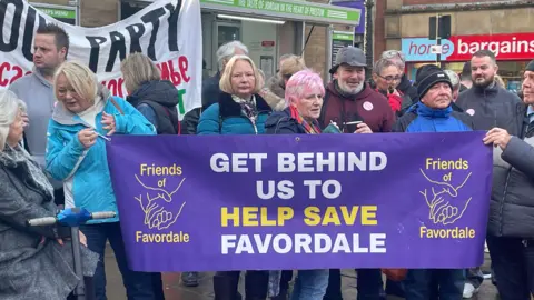 About a dozen people stand behind a purple banner saying "Get behind us to help save Favordale"