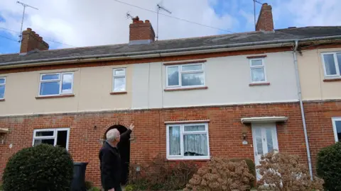 Jean Thompson Barry Thompson pointing at his roof with his back to camera. There are blue skies and white clouds above.