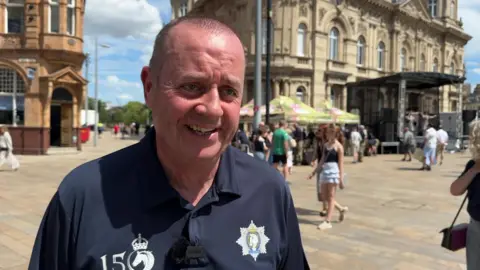 Ian is smiling and wears a dark blue top with regimental logos. Various stalls and buildings can be seen in the background.
