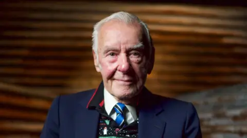 Ted Bush, aged 92, has short white hair and is clean shaven. He is smiling directly at the camera and wearing a navy blue suit jacket over a white shirt, blue tie and patterned sweatshirt.
