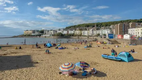 North Somerset Council A sandy beach next to the marine lake in Weston-super-Mare, which is busy with parasols, people sunbathing and swimming in the lake