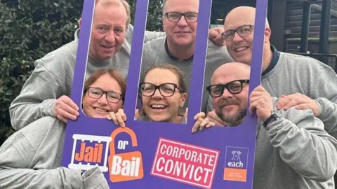 EAST ANGLIA CHILDREN'S HOSPICES Foure men and two women posing behind pretend prison bars for charity picture. Five of the people are wearing glasses. 