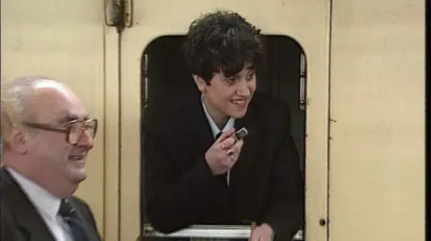 Train conductor Ruth Davies leans out of a yellow box with a silver whistle in hand. He wears a black suit jacket and a white shirt. Next to her sits a bald man in a black and white suit, wearing glasses