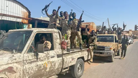 Ken Mungai / BBC Dois caminhões do exército carregando soldados aplaudindo pertencentes às forças armadas sudanesas em uma rua no norte de Cartum - março de 2025