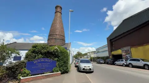 The front entrance to the Moorcroft factory in Stoke-on-Trent. A large blue sign on the side of the road says "Moorcroft". A bottle oven can be seen sticking out of a building in the background.
