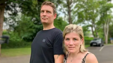 A man with brown hair and a black top stands next to a woman with blonde hair.