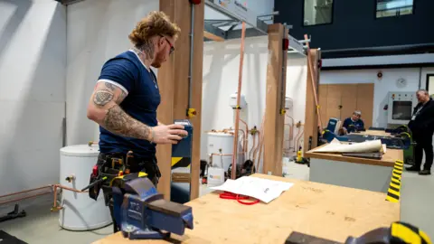 WorldSkills UK A man with ginger hair and a tight blue t shirt is working at a workbench on a gas plumbing project 