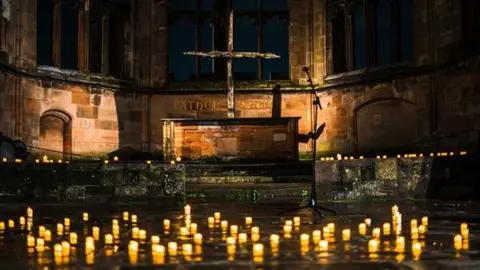 Coventry Cathedral A picture taken from ground level in the cathedral showing small candles on the floor. An old fashioned alter is in the background with a wooden cross behind it. 