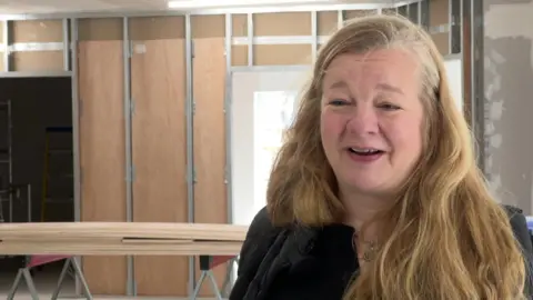Jane Holmes has long blonde hair and is wearing a black top. Behind her is the space being renovated to become the charity's new base.
