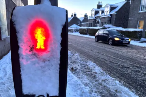 A traffic light 'red man' covered in snow, with a car with its lights on, on a snow-covered road.