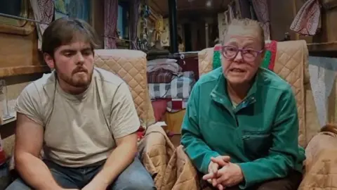 An older woman sits with folded hands next to a man inside a narrow boat