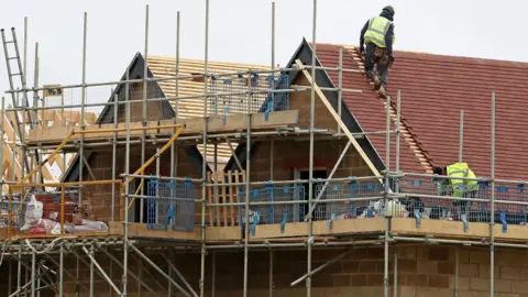 A generic picture of a row of brick houses being built. Scaffolding has been put up around the new builds and there are two builders working on the roof of the properties on the right, closest to the camera.