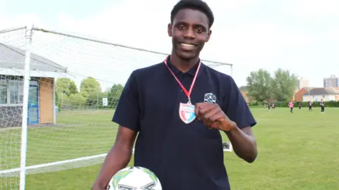 Lawn Manor Academy Bubacarr smiles at the camera while standing on a football pitch. He has a polo top on and holds a football in one arm and a Barcelona badge and lanyard in the other.