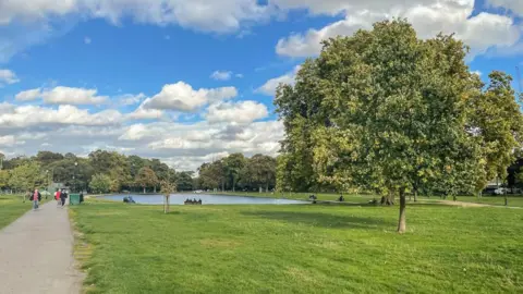 File photo of Clapham Common in the daytime. There is green grass, some trees and people walking on a path and sitting on a bench looking at a pond.