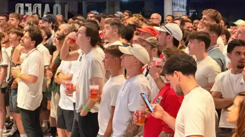 Isobel Gauld A crowd of people wearing England shirts and holding pints of beer look with blank faces towards the screen which is out of shot. One man in looking at his phone.