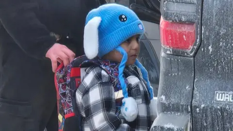 A boy identified as Liam Ramos is shown standing with an immigration agent, in a provided by Columbia Heights Public Schools. Liam wears a blue hat shaped like a bunny, and a plaid grey shirt. A man's hand is seen holding onto his backpack