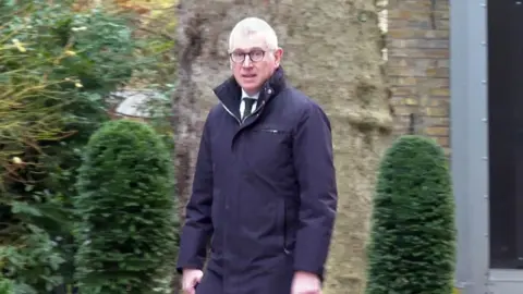 Tim Allan walking in Downing Street wearing a dark coloured coat, a white shirt, dark coloured tie and glasses. There are bushes and a tree in the background.