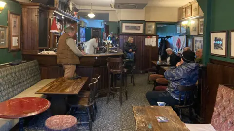 Round and rectangular topped wooden bar tables in the foreground leading to a collection of people enjoying pints of beer standing and sitting around a wooden bar which is to the left side of a long room.