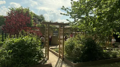A pergola is surrounded by trees and plants with children in the background walking around the memory garde.