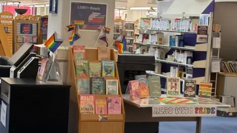 A display of books with several rainbow flags representing the LGBTQ+ community. There is a banner on a table saying "read with pride"