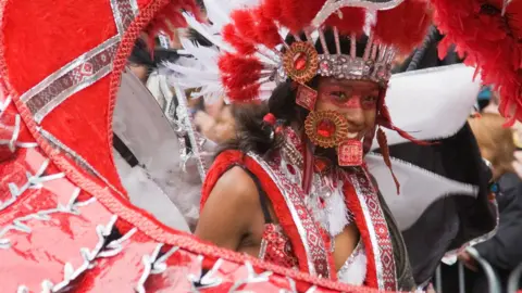 Getty Images A woman with glittery red face paint around her eyes is wearing a large costume that surrounds her with red and white feathers