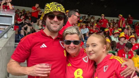 Three people in red Wales tops at a match. On the left a man in a yellow and black bucket hat and sunglasses, and in the middle a woman with short blonde hair and sunglasses. On the right is a woman with reddish hair, her sunglasses resting on her head