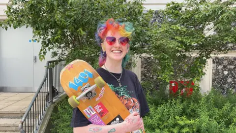 Ozzy Crawley stood with their skateboard in their arms. They have multi-coloured hair and a chain around their neck. They are wearing heart-shaped sunglasses and have piercings on their face and tattoos on their arms.