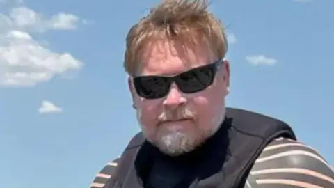 Karl Bushby A man with brown hair and grey beard is wearing a wet suit and sunglasses. Behind him is a blue sky.