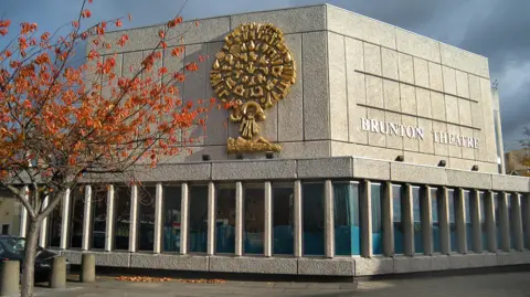 BRUNTON THEATER Side view of the exterior of the Brunton Theatre. The building is gray with a golden design on one side. On the other side are the letters 'Brunton Theatre' in silver. In front of the building, there is a tree with red and orange leaves on three stone pillars.