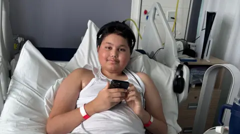 Reece Khan - a boy with short black hair wearing a white vest - sat in a hospital bed. He is holding a phone and is smiling at the camera. 