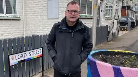 BBC/Andy Mitchell Shaun Pickup stands by the newly decorated Lord Street sign and a huge rainbow-painted street planter.