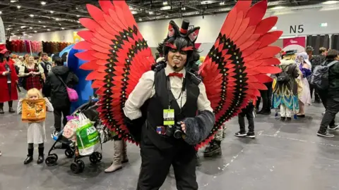 Thomas Copeland / BBC News A fan dressed in a costume consisting of a white shirt, black vest, large red wings and red face paint.