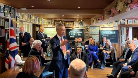 A grey hair man in a dark blue suit, white shirt and red tie is standing in the middle of the room speaking to a group of people. 