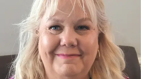 Hudgell Solicitors A woman in her 50s with long blonde hair and blue eyes. She is wearing gold hooped hearings and is smiling.