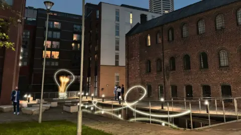 Birmingham Light Festival A large lightbulb, taller than one storey of a building, outside by water, with two buildings being at right angles to the water. A curved line of light goes above the water.