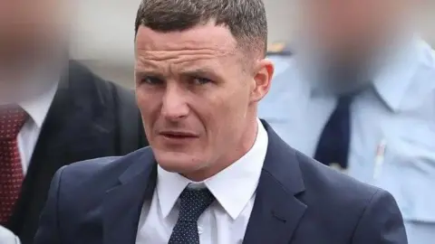 Paddy Cummins Kielan Mooney pictured at a previous court hearing. He has short brown hair and is wearing a suit and tie. Two people, who have their faces blurred, can be seen behind him.