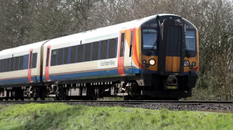 A South Western Railway train makes its way along the railway line near to Basingstoke in Hampshire. Green grass is seen either side and tall trees in the background.