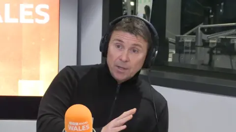 Dean Harris who has dark hair and eyes and is wearing a black zipped up fleece, sat in a radio studio