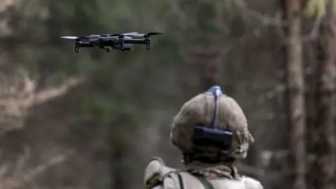 The back of a soldiers head, wearing a helmet as the soldier looks out at a drone flying ahead of them.