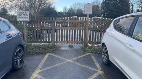 A brown wooden fence, with a white sign saying private car park on the left and a white sign with parking notice on it. On the floor are yellow painted lines to indicate no parking, these were painted to indicate the path. In the background is the Longton Medical Centre carpark