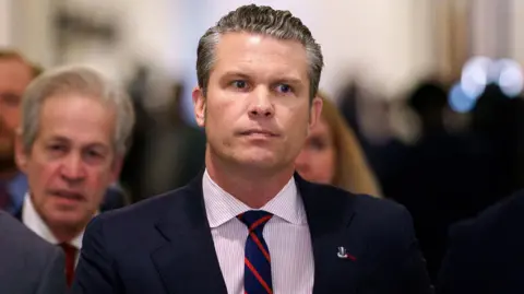 Pete Hegseth is seen up close wearing a blue jacket and striped shirt. He is not smiling and appears to be walking with several people around him blurred in the background