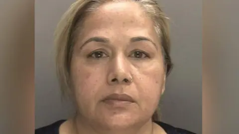Farzana Kauser with blonde hair in a ponytail and a black top, stood in front of a grey background for her mugshot.