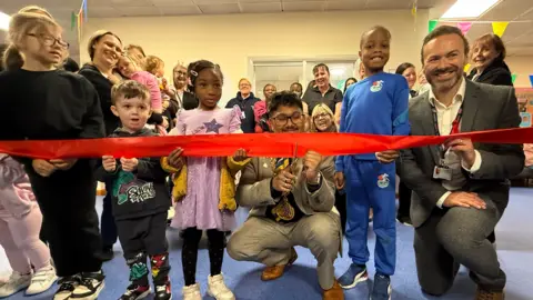 Sunderland Mayor Ehthesham Haque, in the centre of the photo, holds a pair of scissors against a red coloured ribbon. He is wearing a light grey suit and brown coloured shoes. There are numerous children, wearing different outfits, standing next to him and holding the ribbon as he prepares to cut it.