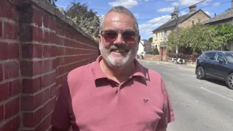 Matt Marvel/BBC A man smiles at the camera as he stands beside a road. He has short grey hair and a grey beard. He wears sunglasses and a red polo shirt. 