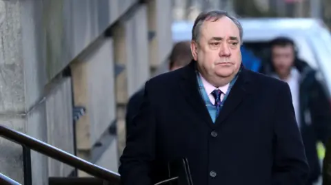 Reuters Alex Salmond walking into court in Edinburgh. He is wearing a dark coat, scarf and pink shirt with blue tie. He is looking to his left and appears in a sombre mood. Two men can be seen in the distance behind him.