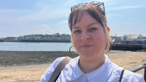 Christine Gray, dressed in a white shirt with a round collar, standing in front of blue skies and a sandy beach near to the Fish Quay in North Shields. 