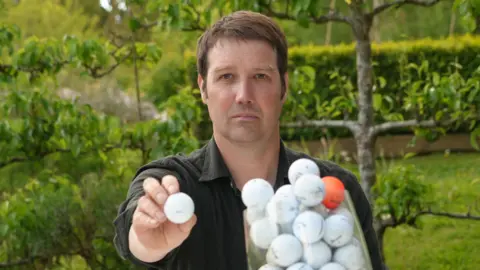 Qays Najm/BBC A head and shoulders image of a man wearing a dark coloured shirt while sitting in a garden with trees behind him. He is holding a white golf ball to the camera while looking into the camera. In front of him is a clear receptacle filled with golf balls - all white ones, except for one orange one.